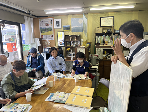 立って話す男性と資料を見ながら座って聞く人達の写真