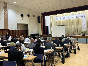 舞台のある広い部屋に並べられた机に大勢の人が着席している写真