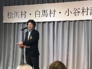 舞台のスタンドマイクの前で話すスーツの男性の写真