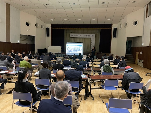 大勢の人が前方のスクリーンを見ながら話を聞いている写真